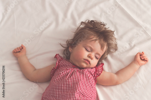 Baby girl sleeping on back with open arms and without pacifier in a bed with white sheets. Peaceful sleeping in a bright room. Pastel retro toned. Soft focus.