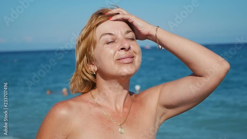  Arty view of a cheerful and stylish blond woman standing, smiling and smartening up on a sandy sea coast with turquoise waters in summer in slow motion photo