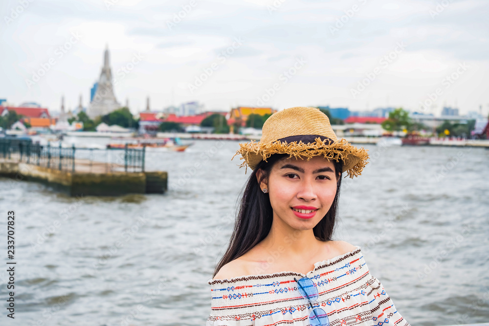 Asian woman tourist wear hat standing smiling with the background is a view of chao phraya river and pagoda arun temple landmark and attractions of Bangkok Thailand.