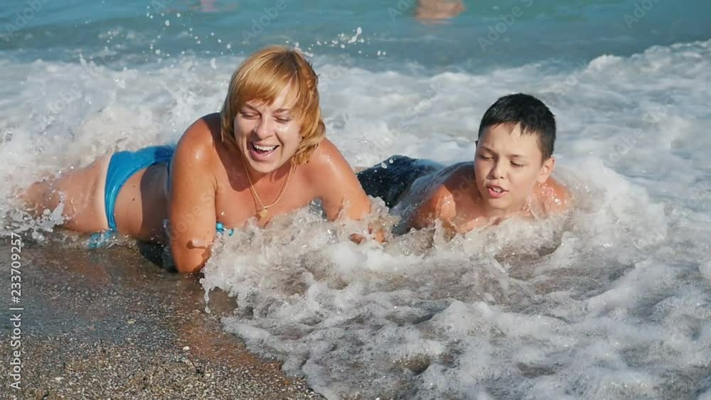 Cheery view of a smiling blond woman and her playing jolly boy lying on their stomachs under tidying waves on sandy beach in summer in slow motion Cheery view of a smiling blond woman and her playing jolly boy lying on their stomachs under tidying waves on sandy beach in summer in slow motion
