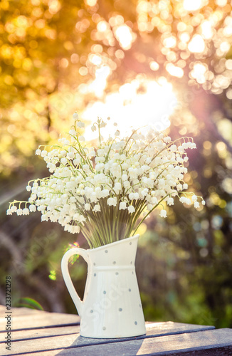 Fototapeta Naklejka Na Ścianę i Meble -  Bouquet of white flowers Lily of the valley (Convallaria majalis) in a white dotted jug shaped vase, outdoors on a table, trees on background beautiful golden hour light.