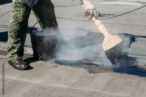 Worker repairs the roof with molten tar from a bucket with a broom.
