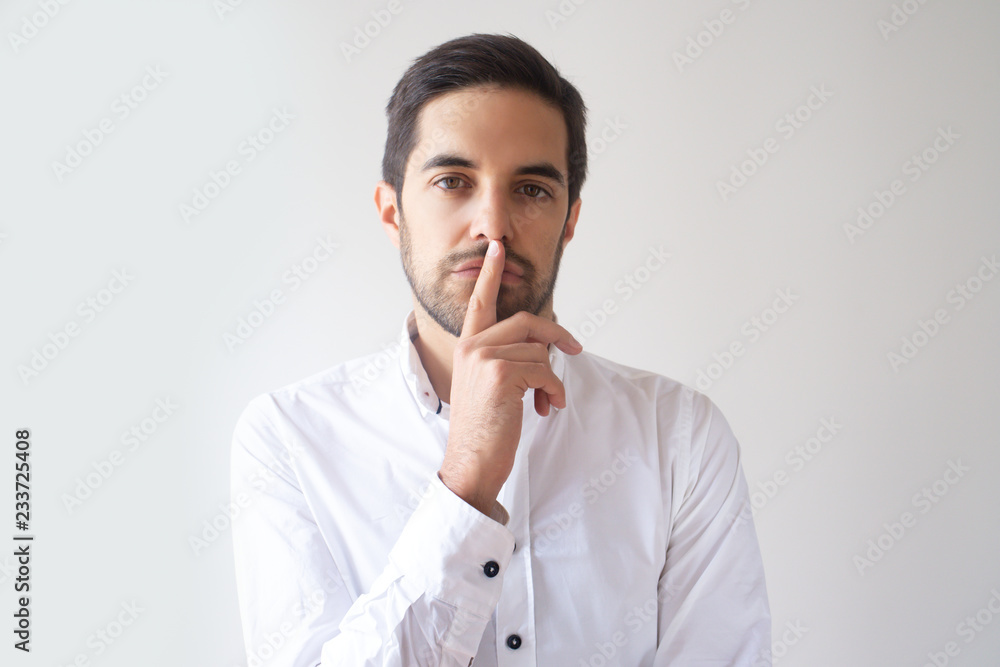 Portrait of young Caucasian man pressing finger to lips. Serious manager in white shirt sharing confidential information. Company spy concept Portrait of young Caucasian man pressing finger to lips. Serious manager in white shirt sharing confidential information. Company spy concept