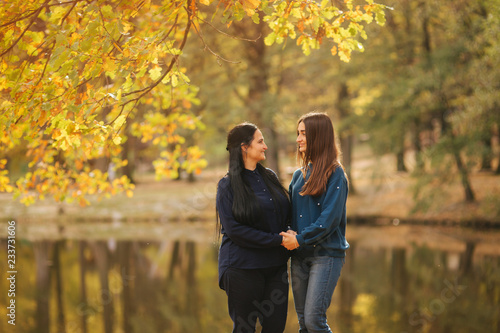 Mother with ldedt daughter stand in front of lake in the park. Yellow tree in autumn time