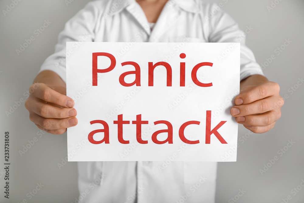 Young man holding sheet of paper with words PANIC ATTACK on light background Young man holding sheet of paper with words PANIC ATTACK on light background