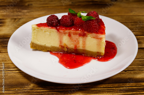 Piece of tasty New York cheesecake with raspberries and raspberry jam in a white plate on wooden table