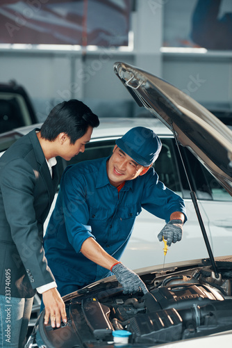 Smiling Asian worker in uniform fixing the car and explaining the operation of the engine to the client in car service