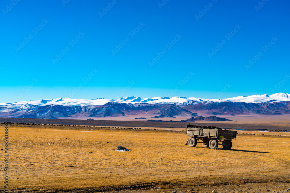 Obraz premium View of beautiful mountain range against the blue sky and the large yellow steppe with a herd of sheep grazing and a trailer loaded with ore near Ulgii in Mongolia