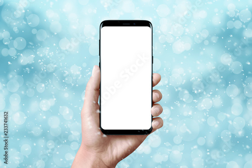 Modern black phone in hand. Isolated, blank screen for mockup. Christmas scene with blue background with snowflakes, light and bokeh.