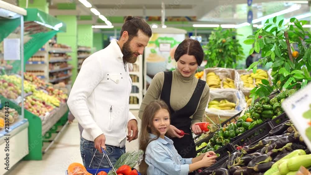 Young happy parents with a child buy vegetables in the supermarket. Mom dad and their little daughter are choosing the peppers and eggplant in a large grocery store. Young happy parents with a child buy vegetables in the supermarket. Mom dad and their little daughter are choosing the peppers and eggplant in a large grocery store.