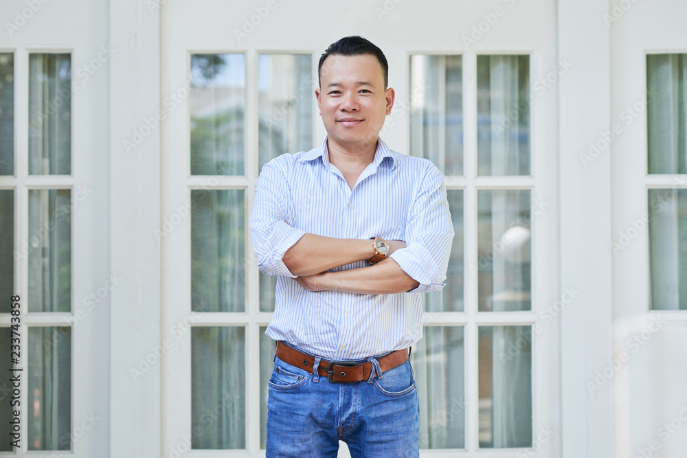 Portrait of happy man standing with arms crossed with his new house with glass doors in the background Portrait of happy man standing with arms crossed with his new house with glass doors in the background