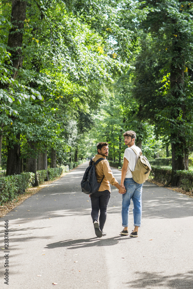 Young gay couple with backpacks walking hand in hand on a road Young gay couple with backpacks walking hand in hand on a road