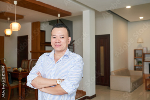 Portrait of Asian successful middle aged man standing with arms crossed in his apartment with modern interior