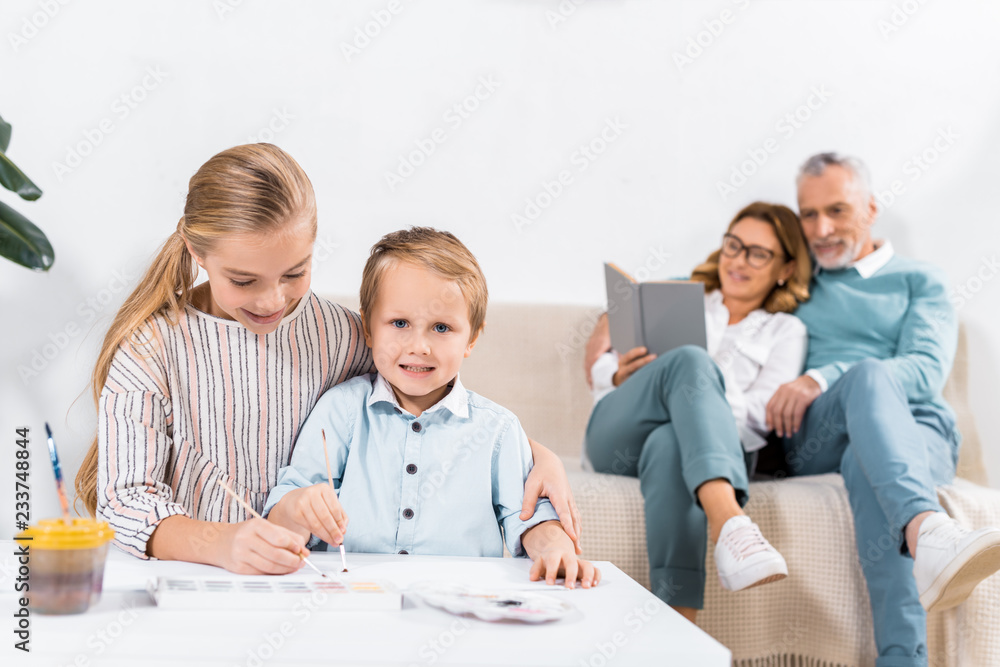 sister and brother painting at table while their grandparents sitting on sofa behind at home sister and brother painting at table while their grandparents sitting on sofa behind at home