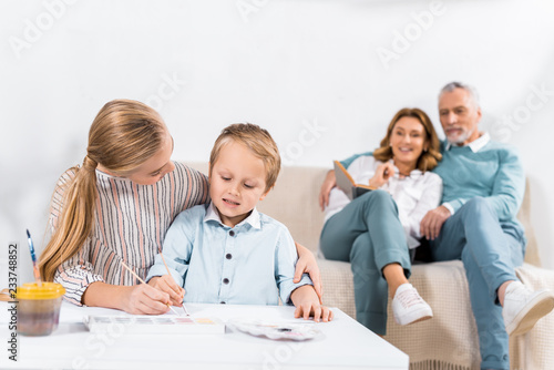 child helping little brother to painting at table while their grandparents sitting on sofa behind at home © LIGHTFIELD STUDIOS