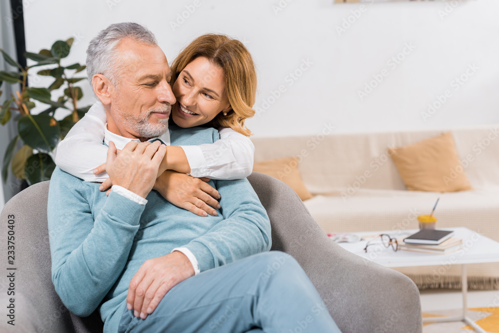 smiling woman embracing middle aged husband in living room at home smiling woman embracing middle aged husband in living room at home
