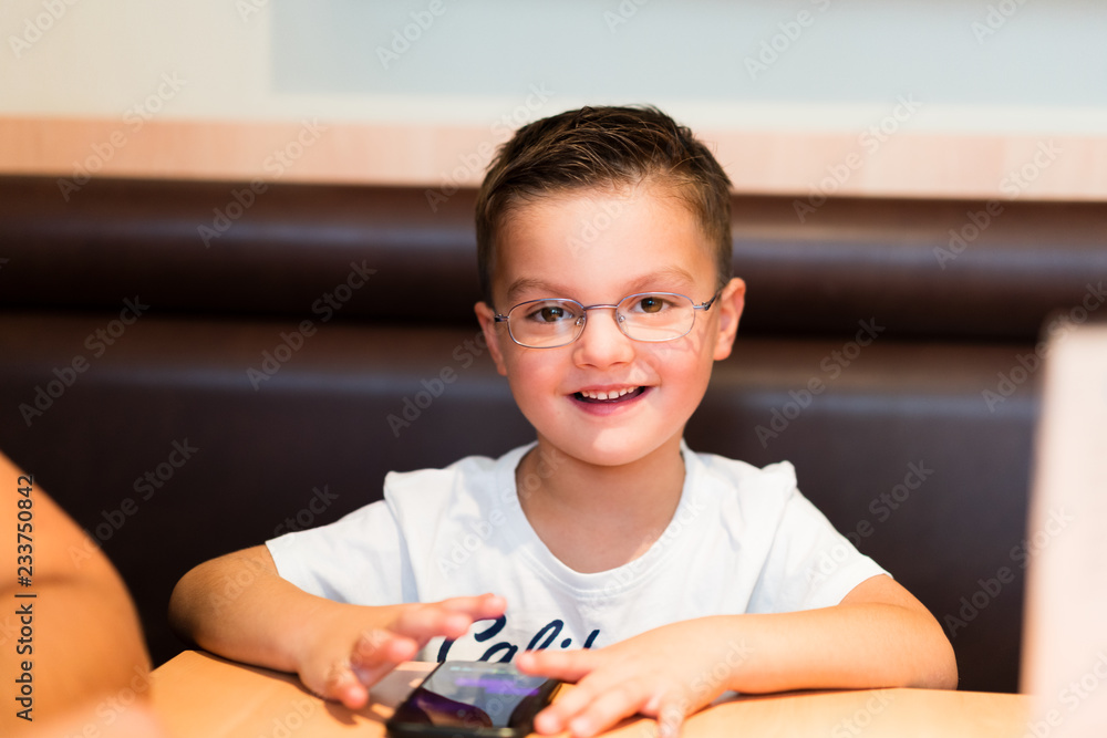 Kind Junge mit Brille spielt mit Handy sitzend am Tisch. Lächelndes Gesicht Kind Junge mit Brille spielt mit Handy sitzend am Tisch. Lächelndes Gesicht