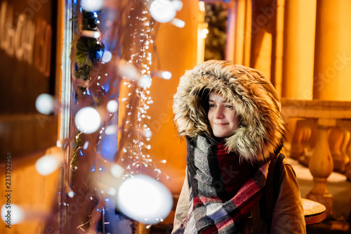 On the street a young girl looks at a shop window decorated with lights and dreams on the eve of Christmas and New Year