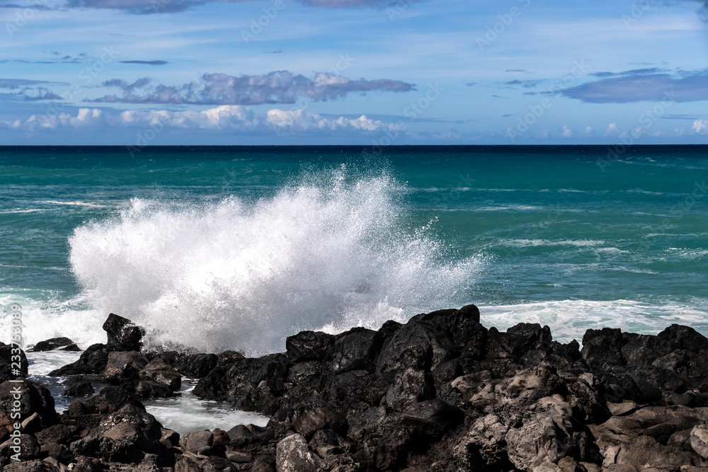 Wave breaking on the western Kona coast of Hawaii's Big Island near South Point. White sea spray thrown into the air; Deep blue-green Pacific ocean, and blue sky with clouds in the background. 