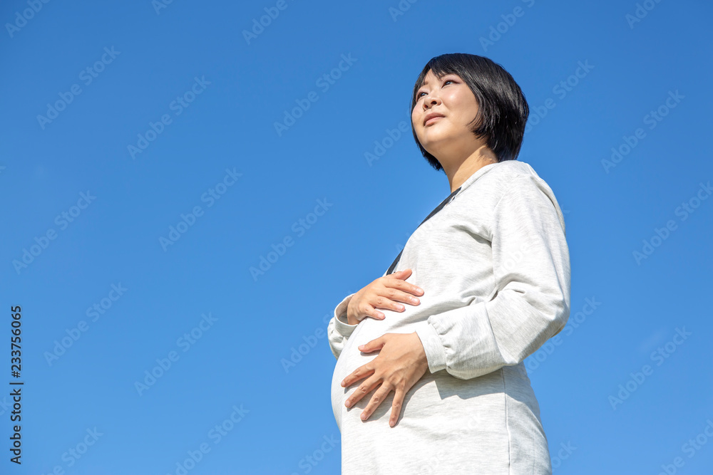 青空を背景に大きなお腹に希望を持って手を当てている臨月の妊婦の様子。妊娠、臨月、愛、楽しみ、幸せイメージ