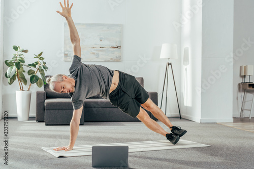 Fototapeta Naklejka Na Ścianę i Meble -  athletic man in sportswear practicing side plank and looking at laptop