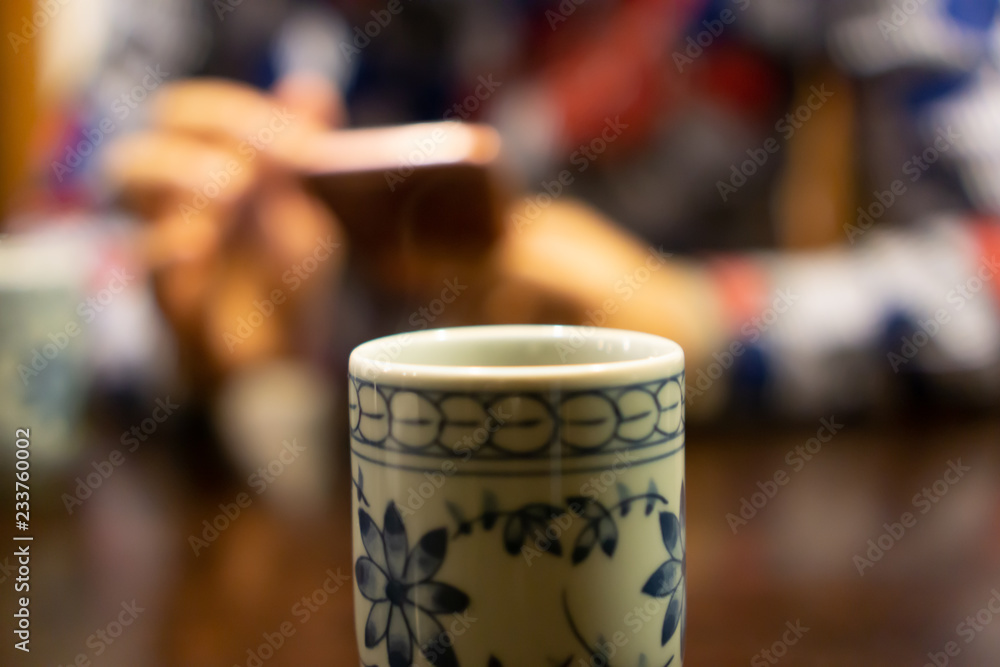 White ceramic cup of Japanese roasted green tea on a table in traditional Japanese restaurant, with young asian woman using smartphone and waiting for her food in the background (The tea is called Hoj White ceramic cup of Japanese roasted green tea on a table in traditional Japanese restaurant, with young asian woman using smartphone and waiting for her food in the background (The tea is called Hoj