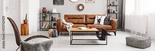 Real photo of brown leather couch with blanket and pillows standing in bright living room interior