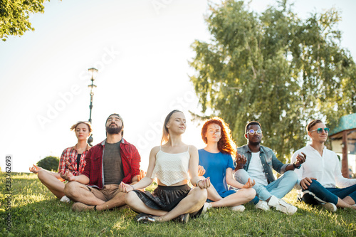 Yoga at park. Interracial friends meditating and spending their leisure time outdoors. Concept of healthy lifestyle, friendship and diversity. Panaromic view of meditating group over nature background