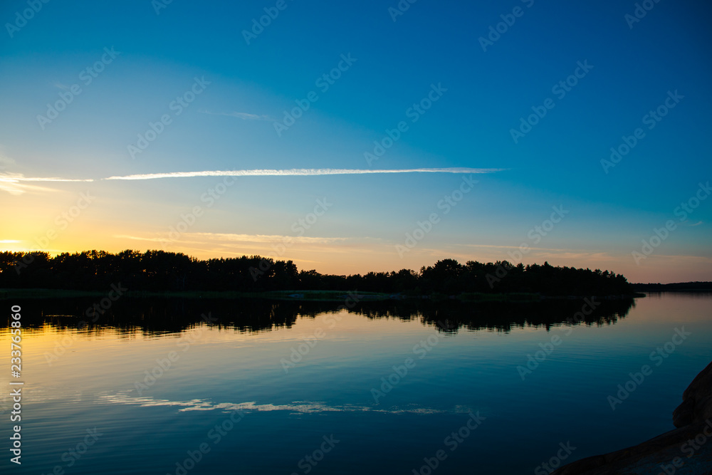 A beautiful and calming sunset accented by a long narrow cloud, a few days after midsummer, reflected on the water of a cove on the Island of Nicklösa in the Åland Islands, Finland.