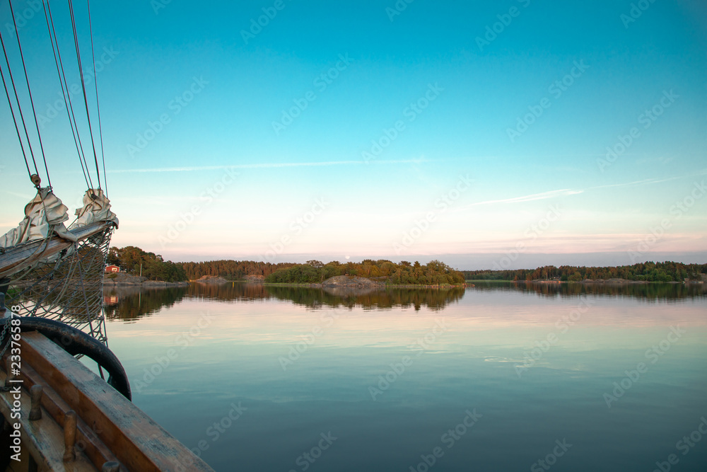 The moon rises softly in the distance, viewed from a three-masted schooner in a cove on the Island of Nicklösa in the Åland Islands, Finland, at sunset a few days after midsummer.