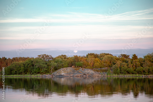 The moon gently rises in the distance behind trees and a rocky shoreline in a cove on the Island of Nickl  sa in the   land Islands  Finland  at sunset a few days after midsummer.