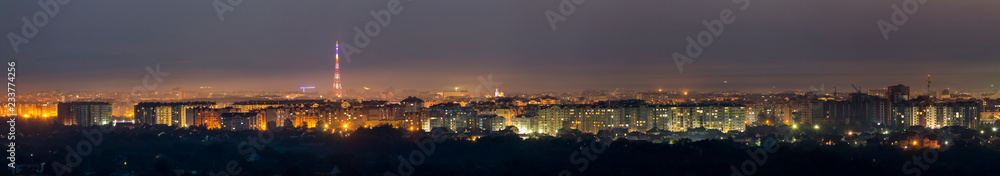 Wide panorama, aerial night view of modern tourist Ivano-Frankivsk city, Ukraine. Scene of bright lights of tall buildings, high television tower and green suburbs on Carpathian mountains background.