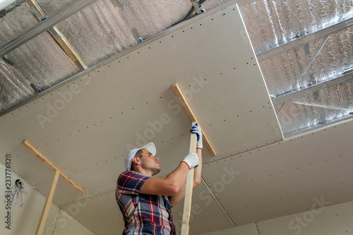 Young worker in protection work gloves fixing wooden holders for drywall suspended ceiling to metal frame insulated with shiny aluminum foil. DIY, do it yourself concept. photo