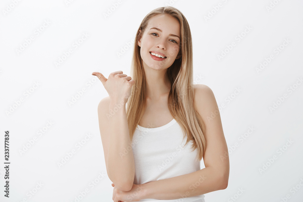 Good-looking friendly and relaxed woman with fair hair tilting head introducing awesome copy space as pointing left with thumb and smiling wanting people check out cool place over gray wall Good-looking friendly and relaxed woman with fair hair tilting head introducing awesome copy space as pointing left with thumb and smiling wanting people check out cool place over gray wall