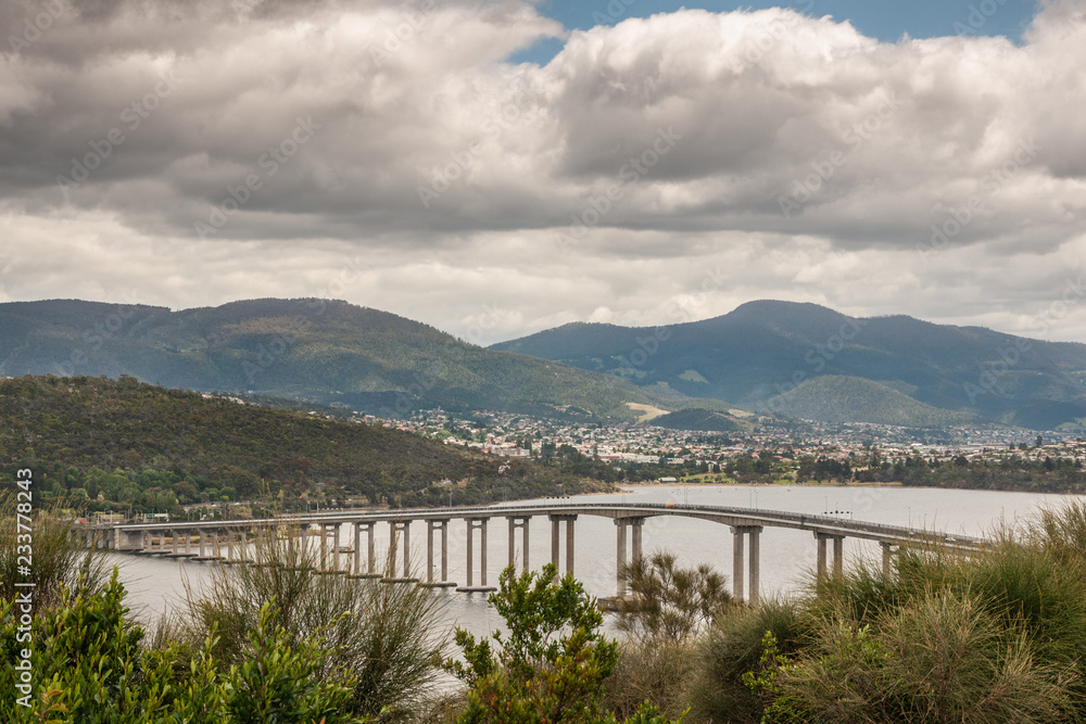 Hobart, Tasmania, Australia - December 13. 2009: Tasman highway bridge over Derwent River with mountains and far off neighborhoods in back under heavy cloudscape. Hobart, Tasmania, Australia - December 13. 2009: Tasman highway bridge over Derwent River with mountains and far off neighborhoods in back under heavy cloudscape.