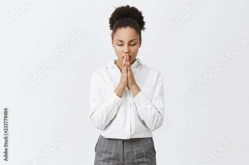 Time to pray and talk with mighty god. Relaxed and focused good-looking dark-skinned female entrepreneur calming down while holding palms together, praying or hoping, being believer in miracles