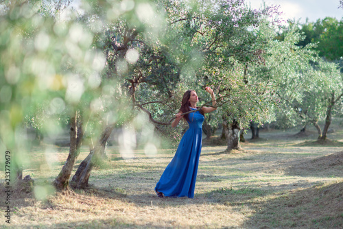 Romantic young beautiful girl with long fair hair in blue dress walking among olive trees in garden. Attractive woman dreaming olive's grove in Tuscany, Italy, Europe, soft focus.