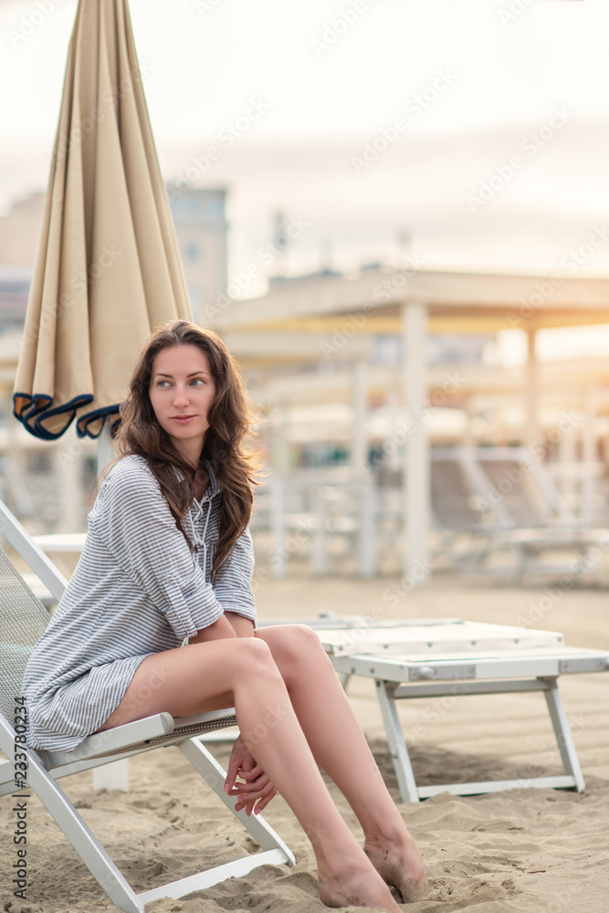 Romantic young beautiful girl with long fair hair in short dress sitting in chaise-longue on the beach. Attractive sleepy calm woman dreaming at dawn on the shore in Italy, Europe, soft focus Romantic young beautiful girl with long fair hair in short dress sitting in chaise-longue on the beach. Attractive sleepy calm woman dreaming at dawn on the shore in Italy, Europe, soft focus