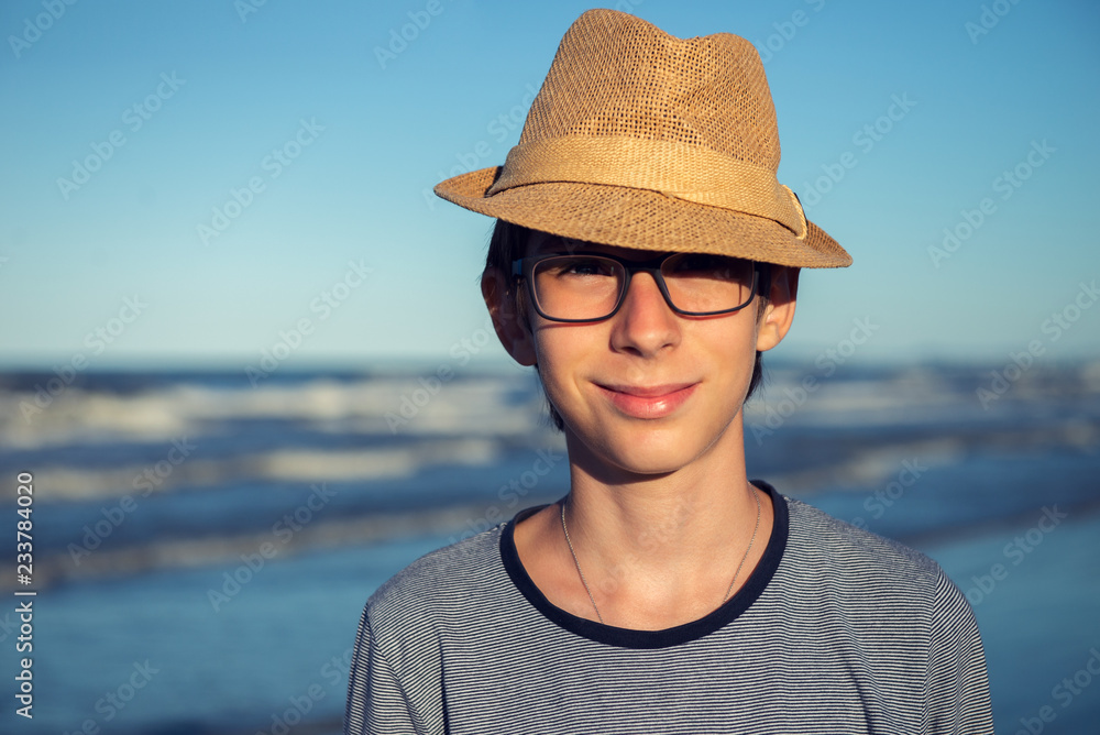 Young boy posing in hat at the summer beach. Cute spectacled smiling happy 12 years old boy at seaside, looking at camera. Kid's outdoor portrait over seaside, fashion hipster style.