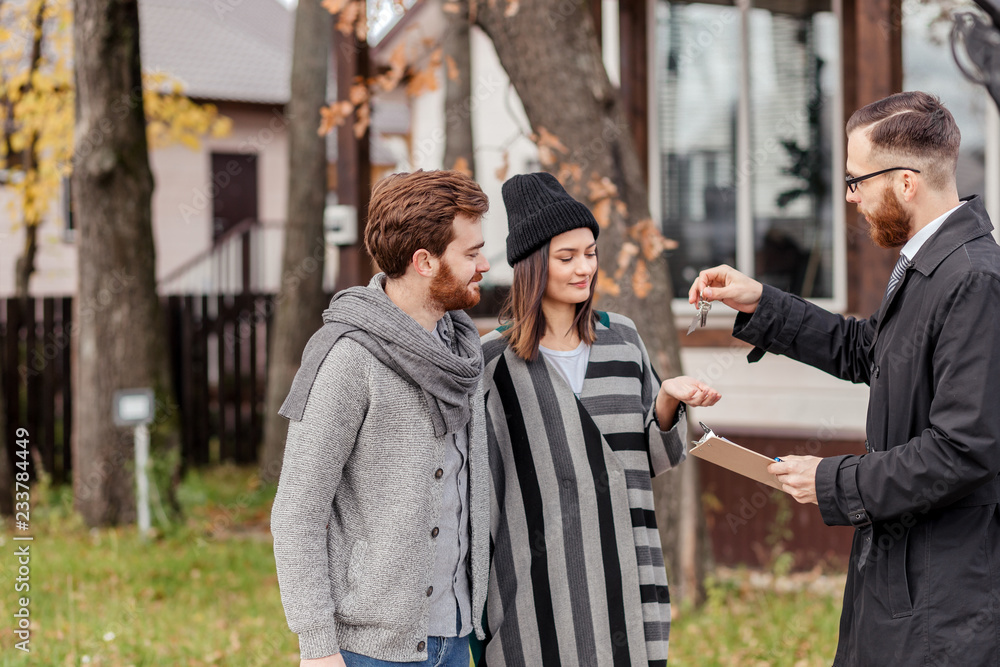 Handsome confidently looking realtor in classic suit and cloak is giving key to the new apartment to happy young couple, standing outdoors, near the house