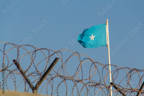 Canvas Print The Somali flag of Somalia, light blue with a five-sided white star, flies on a flag pole above a wall with barbed wire or razor wire in Mogadishu, the capital of Somalia