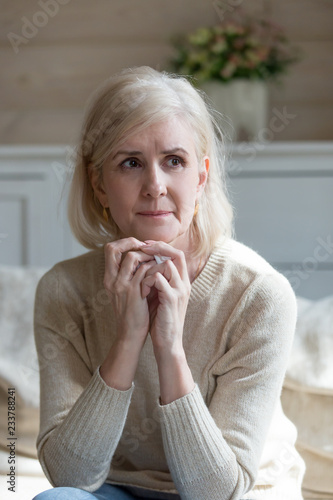 Upset old lady crying sitting on couch alone at home, sad aged woman looking in distance thinking about passed husband missing him, thoughtful senior female remembering good times feeling lonely photo