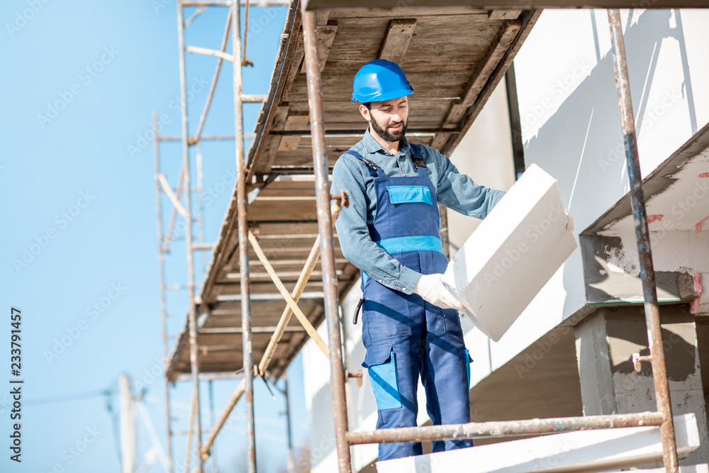 Builder warming a building facade with foam panels standing on the scaffoldings on the construction site
