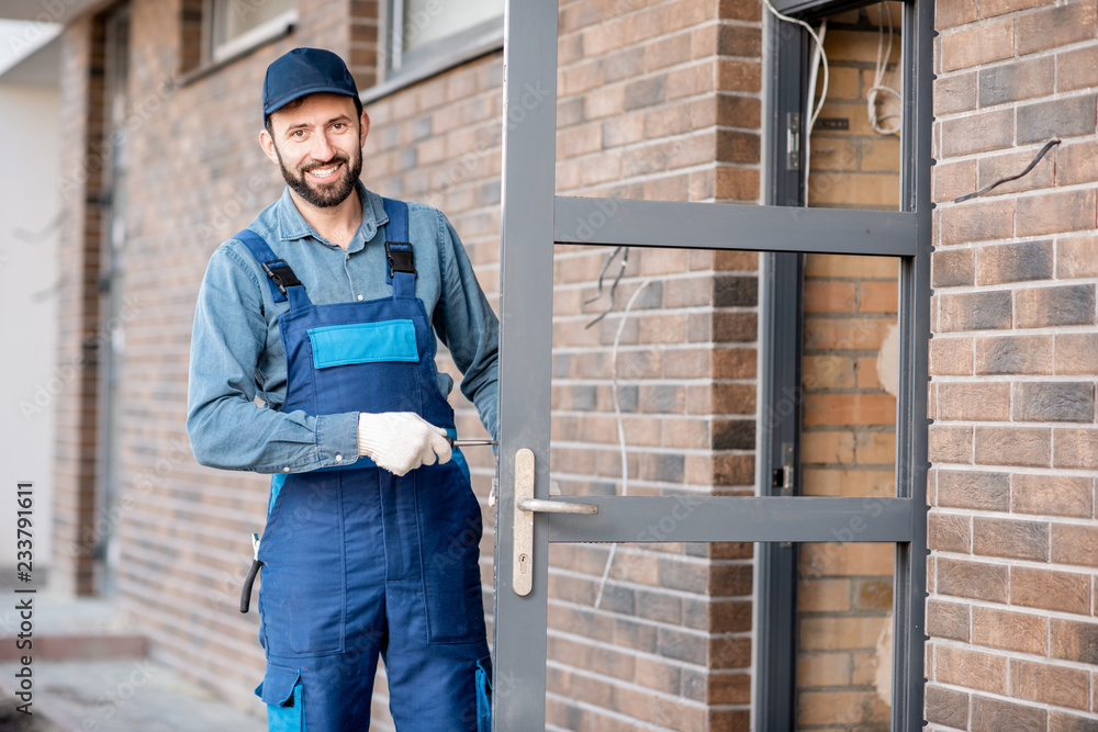 Builder in uniform installing a door lock into the entrance door of a new house outdoors Builder in uniform installing a door lock into the entrance door of a new house outdoors