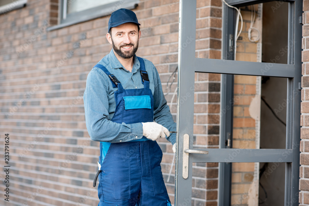 Builder in uniform installing a door lock into the entrance door of a new house outdoors Builder in uniform installing a door lock into the entrance door of a new house outdoors