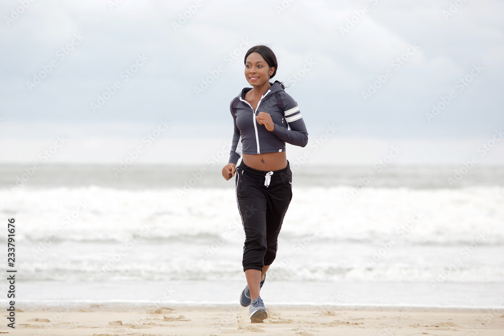 Full body sporty young black woman running on beach by water