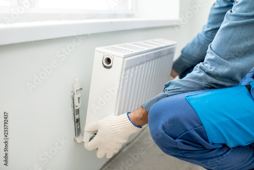 Workman mounting water heating radiator on the white wall indoors, close-up view photo