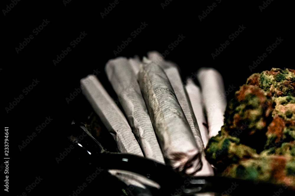 A handful of joints in the end of a glass dish next to a pile of marijuana bud isolated in black.