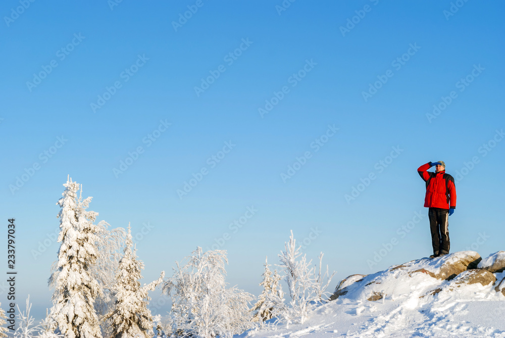 hiker man in winter sportswear looking into the distance standing on a cliff against the blue sky over a winter snow forest