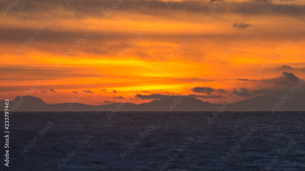 Brilliant orange and yellow sunset at sea with mountains and clouds in silhouette